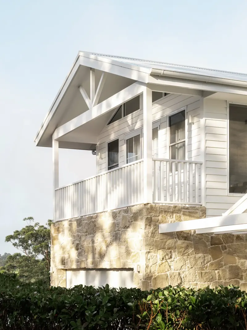 A white weatherboard house with a gabled porch, set on a stone base behind green hedges
