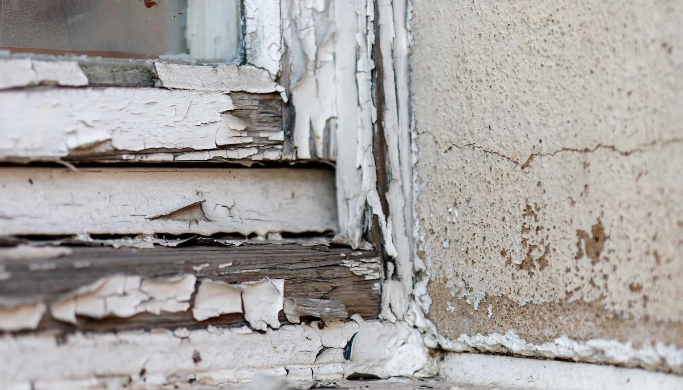 Cracked white paint peeling off a weathered window frame and adjacent concrete wall.